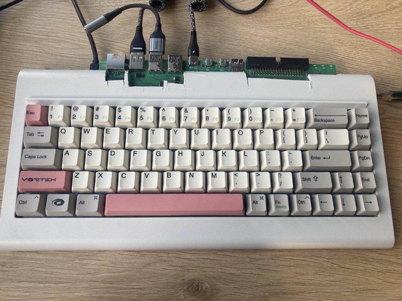 Photo of a mechanical keyboard with a circuitry and wires sticking out of the back, standing on a wooden table. The keyboard is beige with some light-red keys (one of them reads 'Vortex'.) the circuit in the back has several USB ports, Ethernet port, GPIO pins, HDMI and Type C ports. There are several cables plugged into USB ports. The place the circuit is plugged in looks janky and cut out by hand.